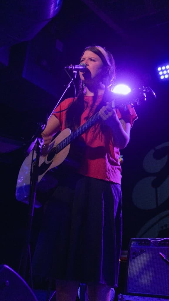 Performer singing into a microphone with an acoustic guitar under purple stage light
