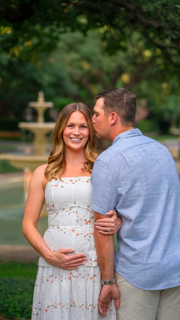 Man kissing an expectant woman on the temple in a sunlit park