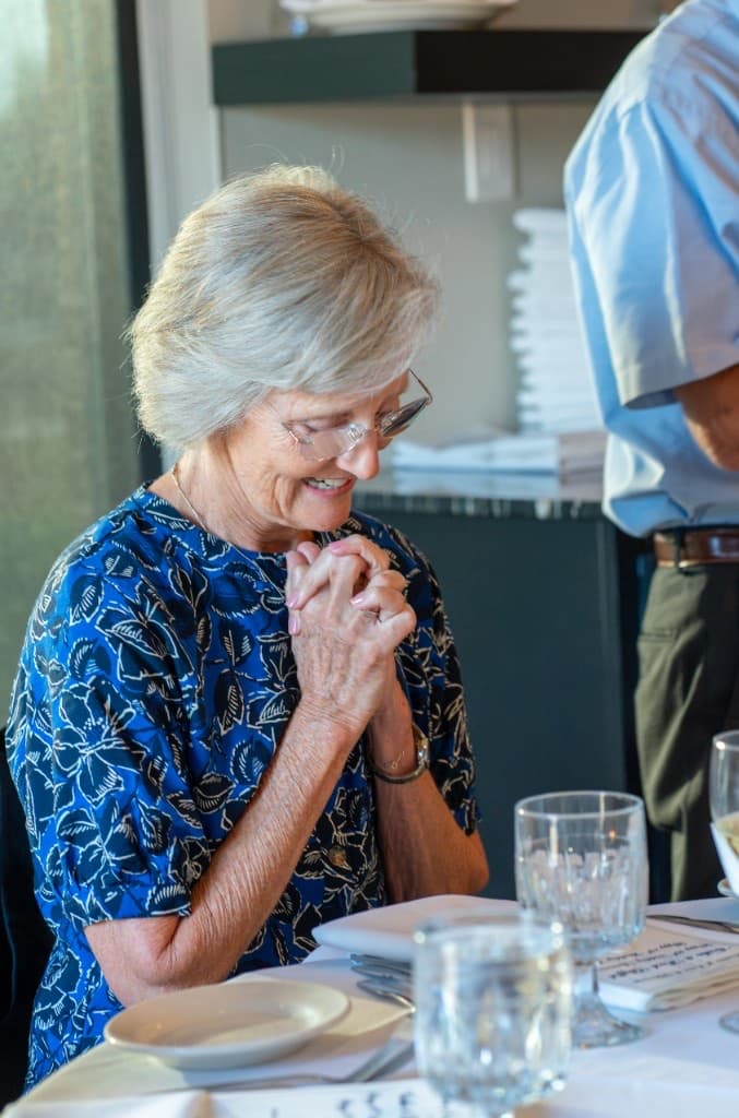 Elderly woman with hands clasped, smiling at a set table